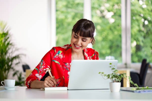Happy young woman wearing red floral pattern jacket sitting at the desk over laptop in the creative workplace.